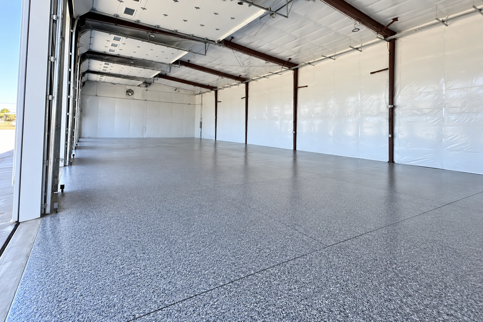 A large industrial garage has a smooth gray speckled floor coating and white walls with exposed steel beams.