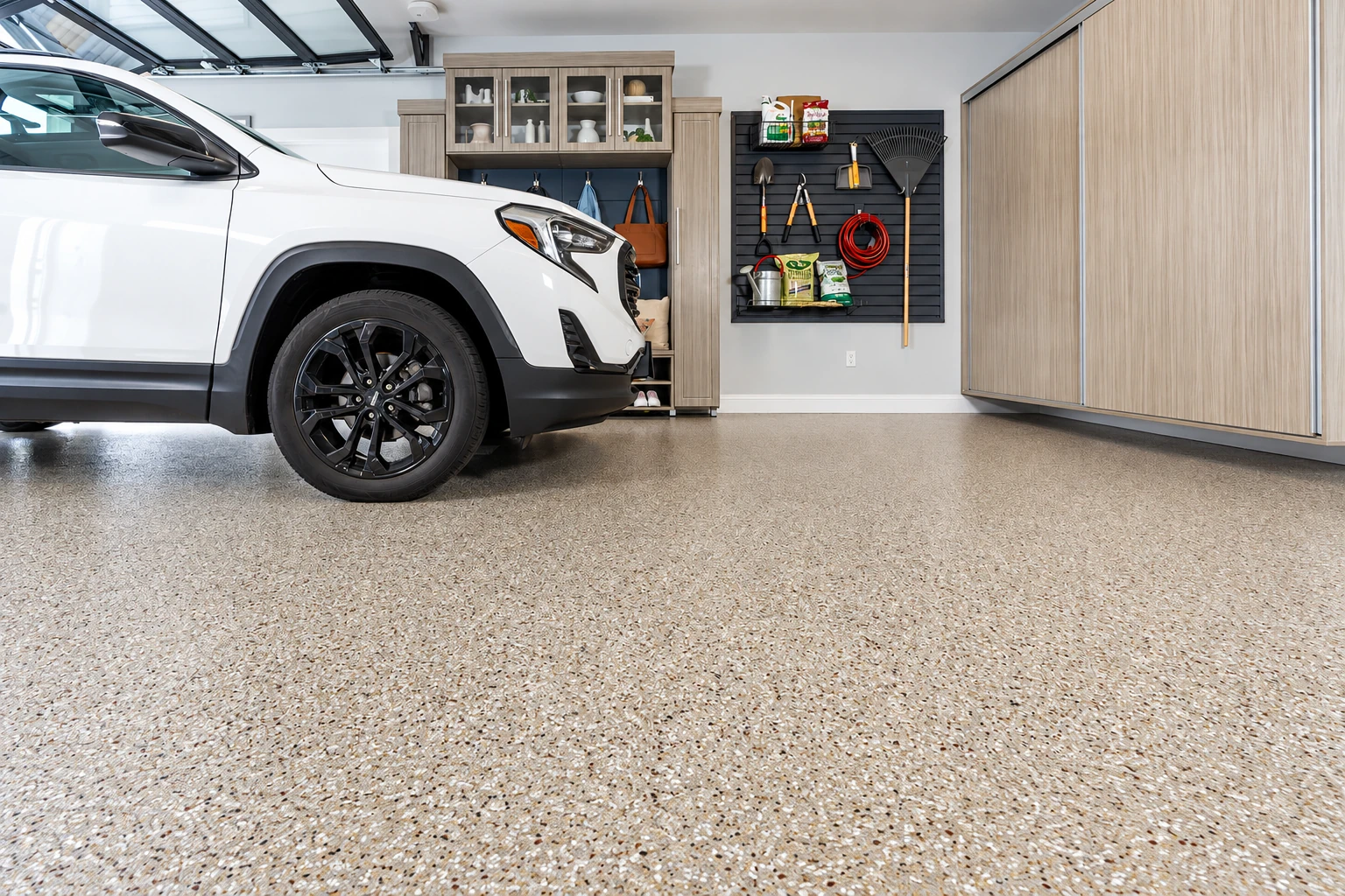 A white SUV sits in a tidy garage with a tan-speckled coated concrete floor and wood cabinets.