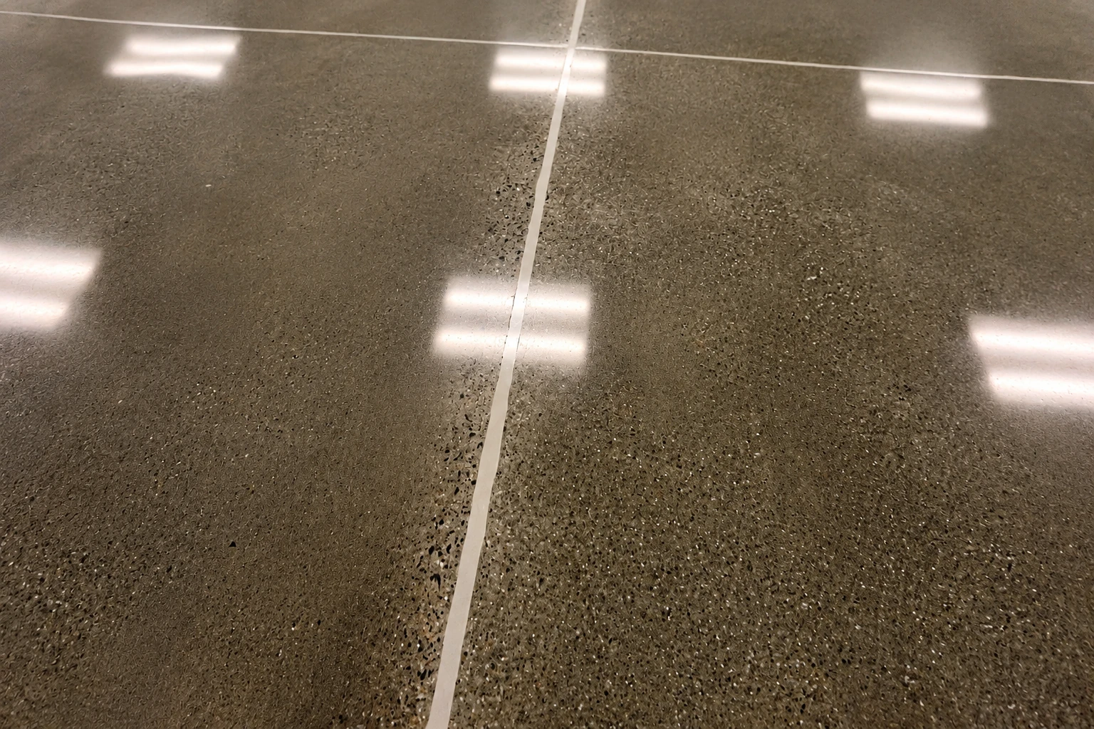 Close-up of polished dark concrete flooring with a narrow pale joint and reflected ceiling lights.